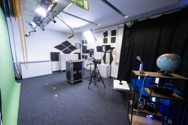 Blick in einen kleinen Studioraum. Links an der Wand ein Greenscreen. Darüber an der Decke befestigt eine Studioausleuchtung. Gegenüber sind zwei Kameras auf Stativen. Rechts ein schwarzer Vorhang der sich hinter einer blauen Couch befindet.